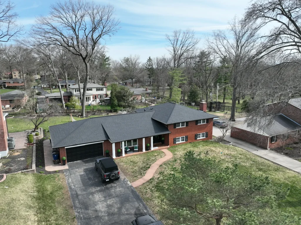 Aerial view of a brick home in Grantwood Village with a new dark gray roof, black garage door, and large trees surrounding the property.