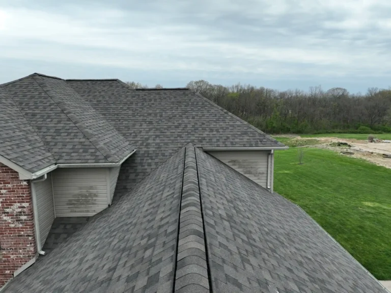 A view of a roof from above showing asphalt shingles with a cloudy sky and a green lawn in the background.