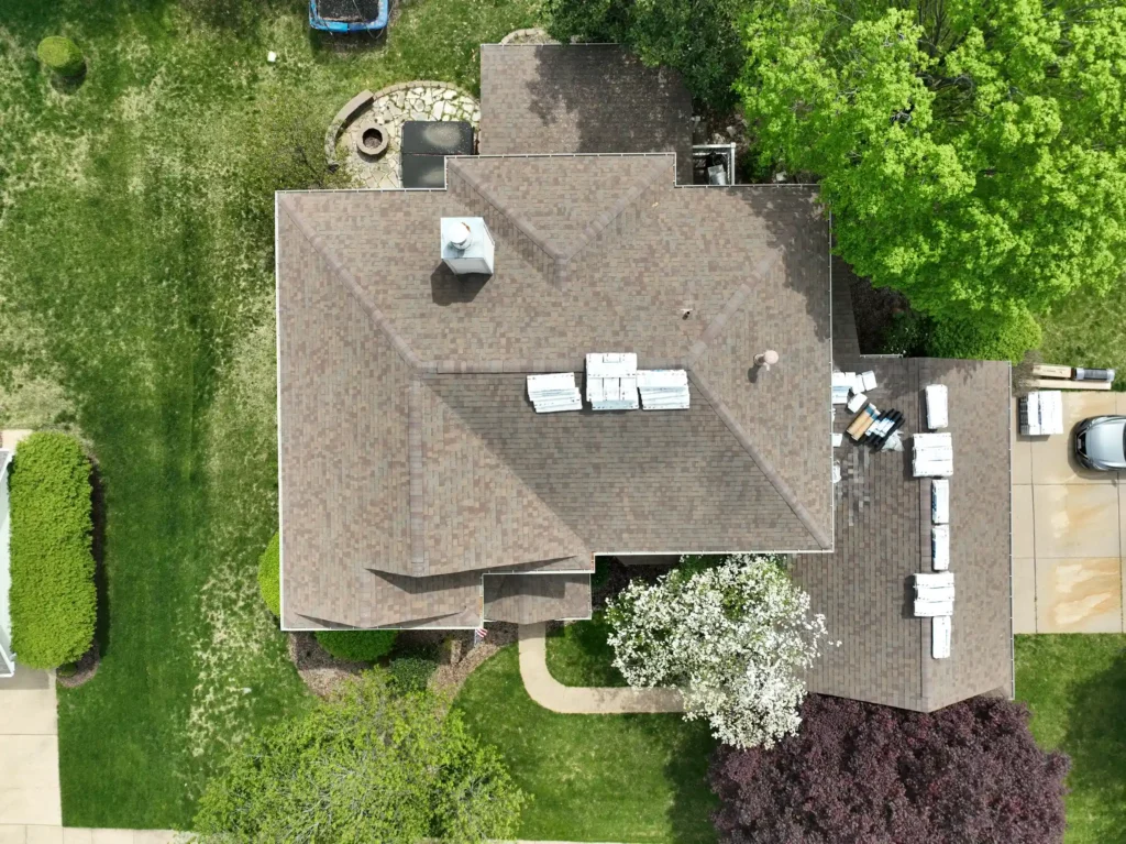 Aerial view of a house with a newly installed roof and stacks of roofing materials on top, surrounded by green trees and lawn in Lake St. Louis.